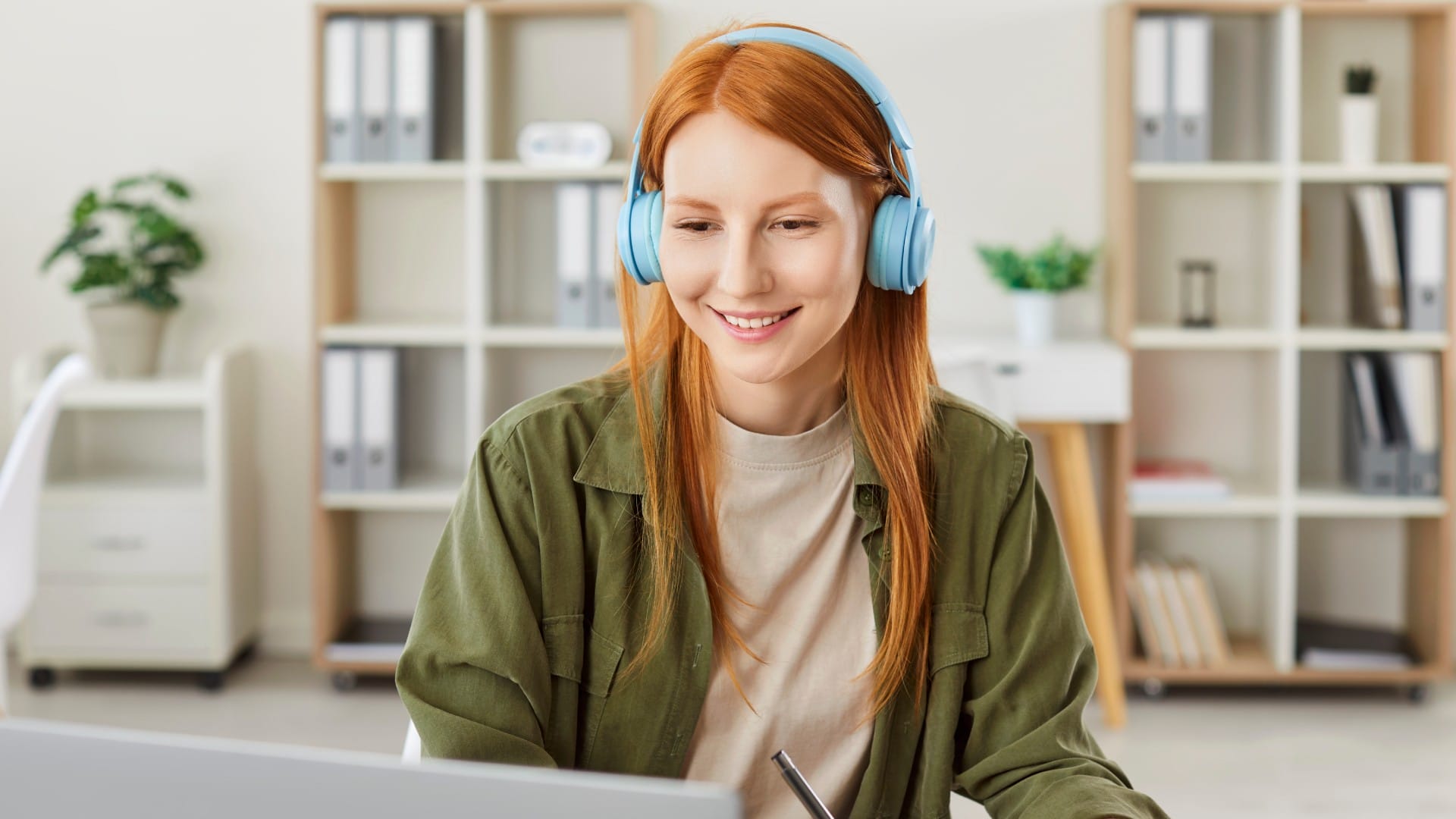 Persona sonriente con cabello rojo con auriculares azules y una chaqueta verde, sentada en un escritorio. El fondo muestra estanterías y una planta de interior, transmitiendo un ambiente de trabajo tranquilo y enfocado.